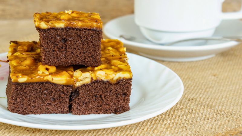 Rich chocolate brownies with a crunchy nut topping are presented on a white plate next to a steaming cup of coffee.
