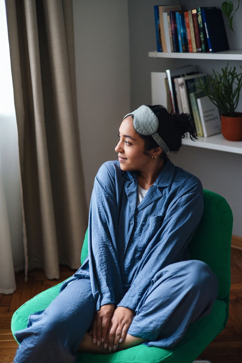 A woman sits comfortably in a green chair by a window, wearing blue pajamas and a sleep mask, enjoying a peaceful, private moment at home with a bookshelf in the background.