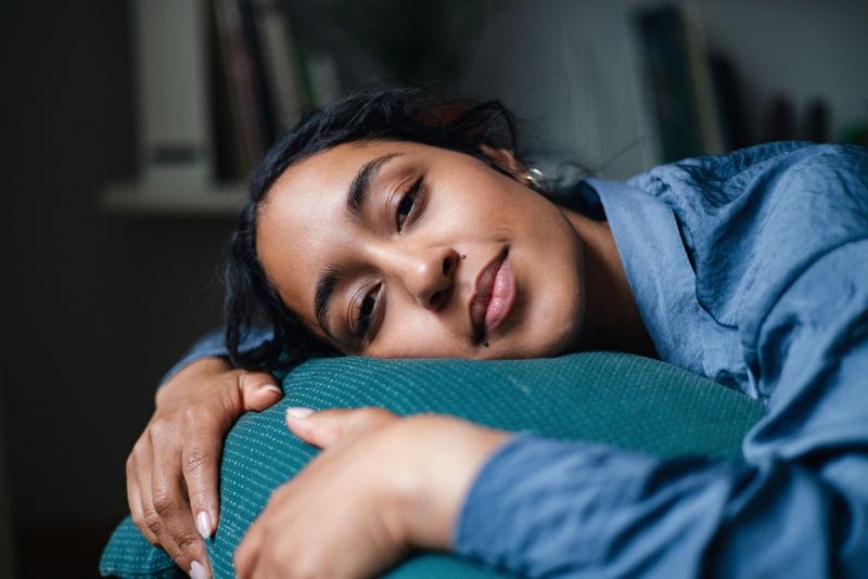 A woman lies on a teal cushion in a cozy home setting, exuding calm and comfort. The soft lighting and relaxed pose capture a quiet, everyday lifestyle moment of rest and ease.