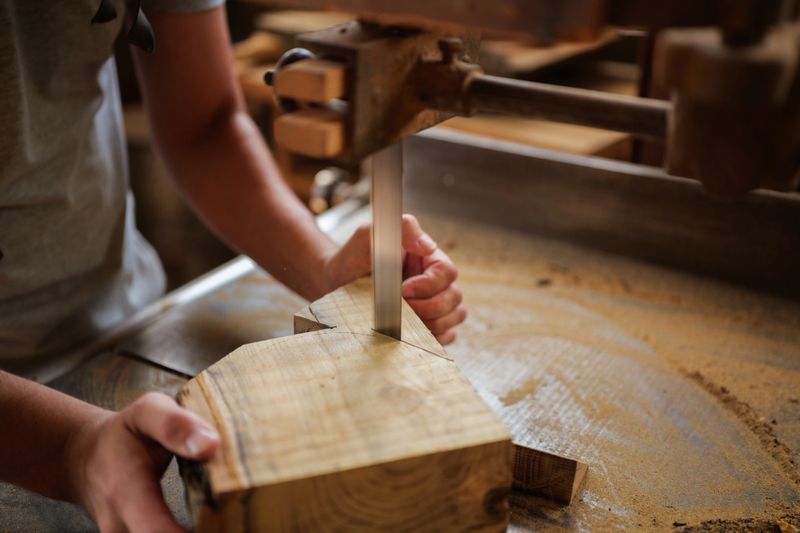 Carpenter carefully guiding a piece of wood through a vertical bandsaw, creating precise cuts in his old workshop, showcasing traditional handwork techniques