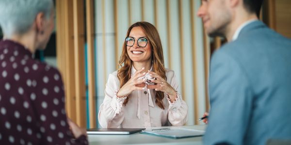 A woman in glasses smiling and talking with two colleagues in a meeting.