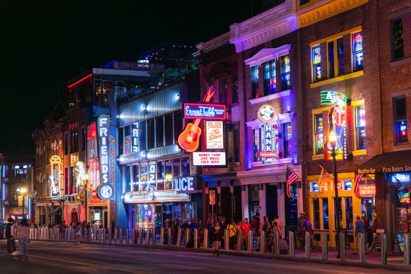 Aerial night view of Nashville's famous Broadway entertainment district in downtown Nashville, Tennessee. The vibrant neon-lit honky-tonk bars and music venues line the historic street, including Ernest Tubb Record Shop with its iconic guitar sign. The colorful LED lighting illuminates the brick buildings that house legendary country music venues, bars, and restaurants. Crowds of visitors can be seen walking along the sidewalks between the establishments. This entertainment corridor is the heart of Nashville's music scene, featuring live country music venues, rooftop bars, and tourist attractions. The area represents Nashville's reputation as Music City USA, with its concentration of historic music venues and modern entertainment establishments.