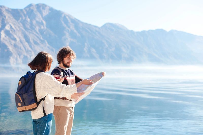 Couple travelers using map at winter sea beach. Happy tourists with backpacks by blue mountains. Young adults man and woman enjoying travel, vacation, adventure. Lifestyle moment. Copy space.