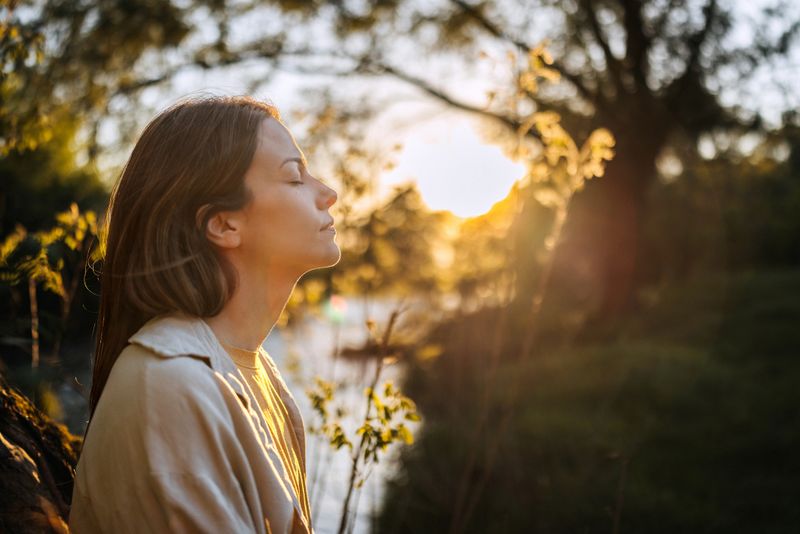 Young woman with long hair enjoying sun with closed eyes getting natural vitamin D outdoors. Peace of mind. Mindfulness, mental health, spirituality, well-being, unwind yourself