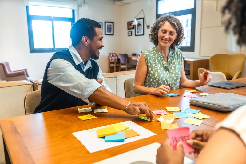 Colleagues of mixed ages brainstorm and share ideas around a table in a bright modern coworking office, using sticky notes and laptops for collaborative planning and innovation