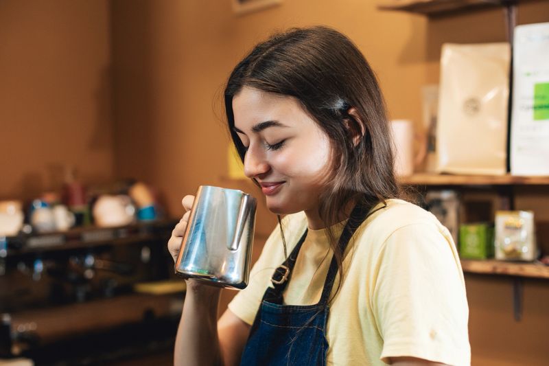 A young barista stands at the counter holding a metal pitcher close to her face, smelling the aroma before making a coffee drink. She wears an apron and works in a cozy café environment.