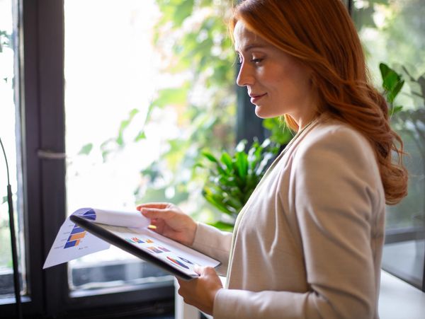 A woman reviewing colorful charts and graphs on paper and tablet by a window.
