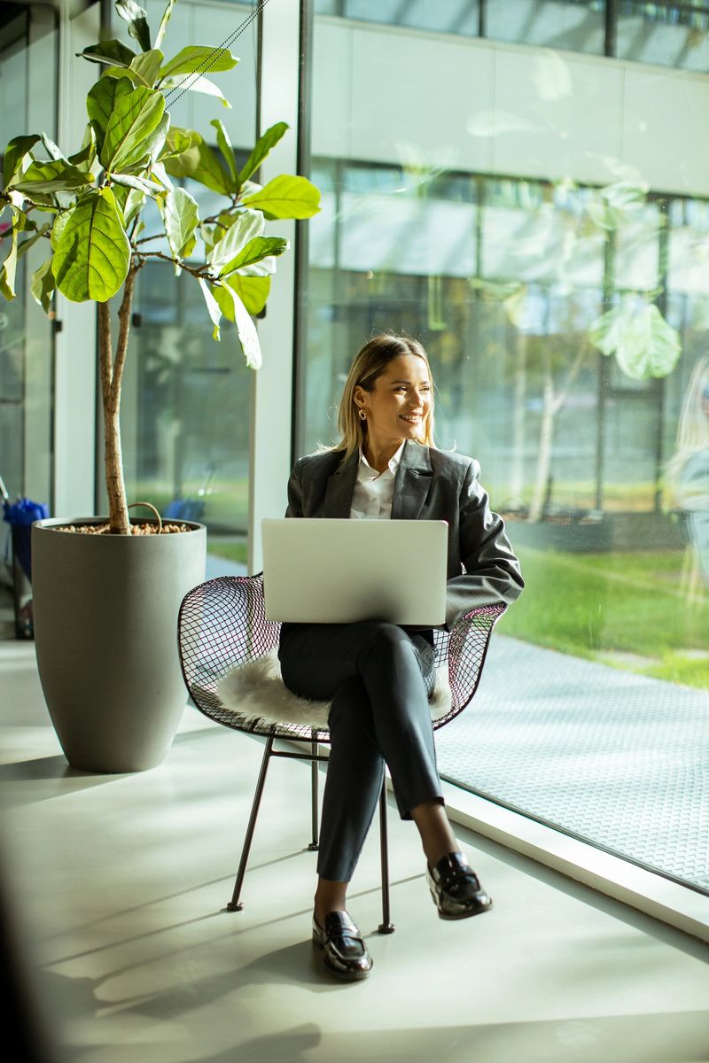 A woman is seated in a modern chair, smiling while using her laptop. She is in a spacious office with greenery and sunlight streaming in through large windows.