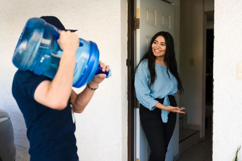 Water delivery service employee bringing a full jug to a happy woman at her home, valuing comfort and convenience