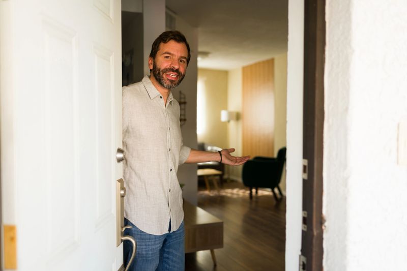 Smiling man welcoming people to his house, standing in the doorway with an open hand gesture, offering hospitality