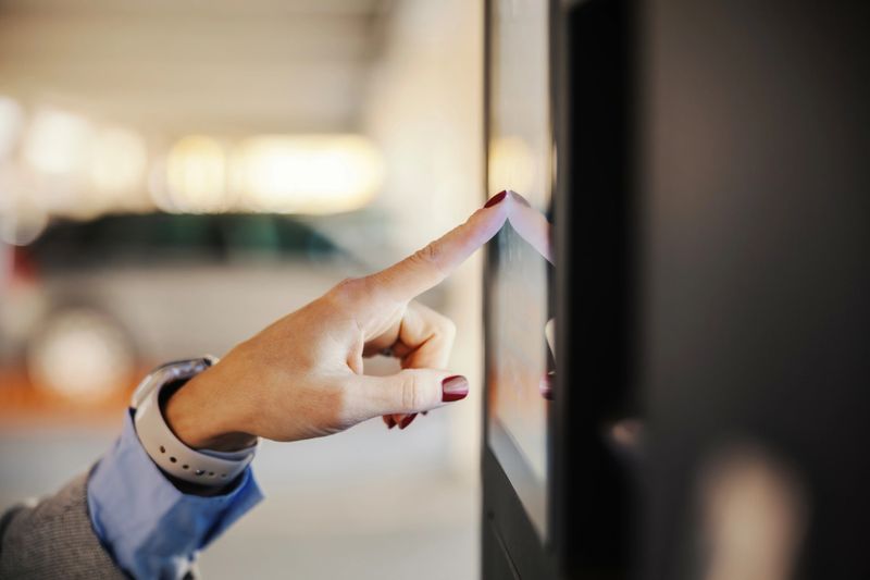 Woman's hand with red nail polish touching a public touchscreen kiosk, interacting with modern technology in a parking garage