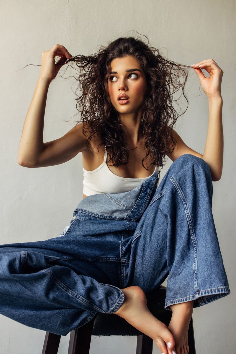 A stylish studio portrait featuring a young woman with voluminous curly hair, wearing a white tank top and oversized denim jeans. She poses with hands in her hair, creating a relaxed, confident, and fashionable vibe suitable for lifestyle, beauty, and fashion campaigns.
