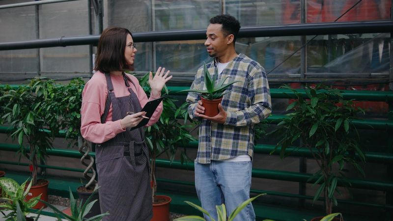 A woman with a tablet engages in conversation with a man holding a potted plant in a vibrant greenhouse surrounded by lush greenery