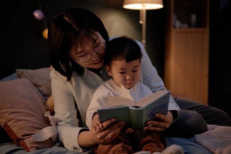 Asian middle aged woman sitting with Asian child reading book together in evening at home, woman holding book while child looking at pages, warm family interaction on bed