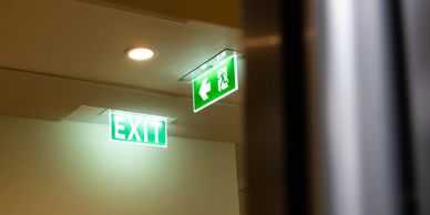 Illuminated green exit signs above a partially open door in a dimly lit corridor.