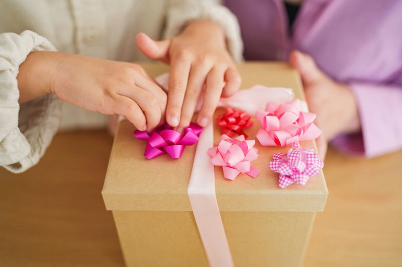 Close-up view of hands decorating a gift box with pink and purple ribbon bows. Warm family moment representing care, creativity, and preparing a special present.