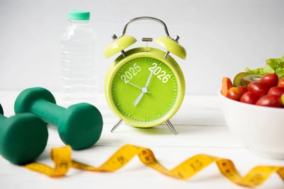 Green alarm clock with 2025 and 2026, dumbbells, water bottle, salad, and measuring tape for fitness.