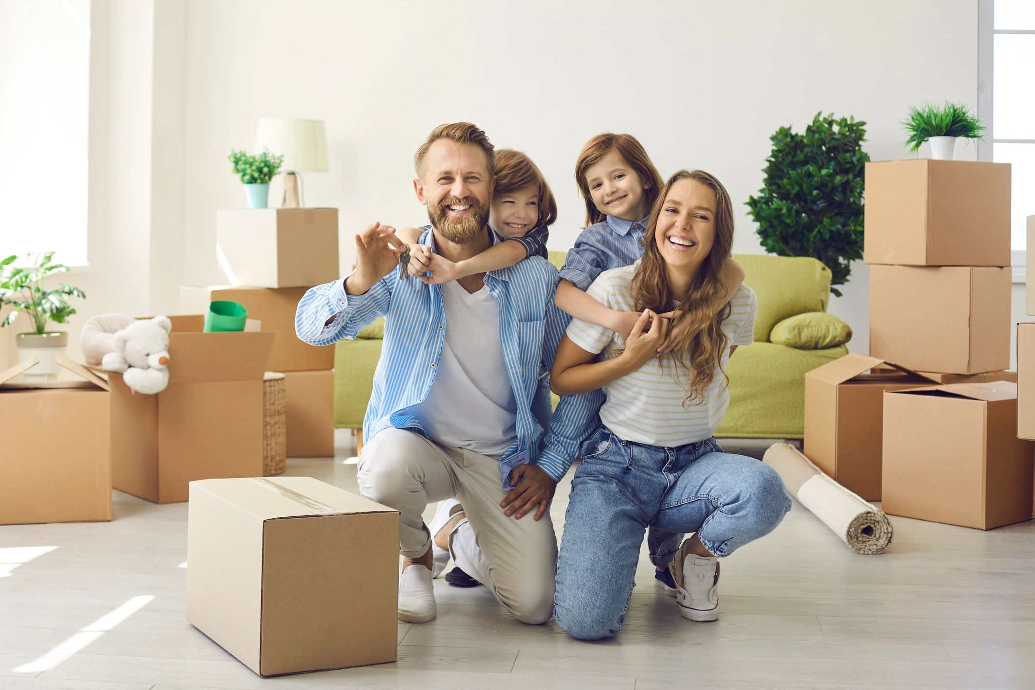A happy family of four smiling on the living room floor of their new home, with the father holding u