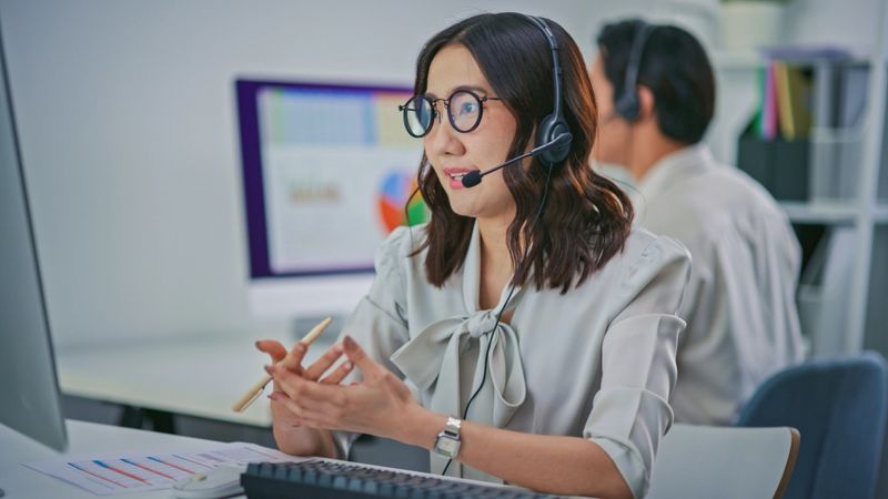 A professional Asian female call center agent wearing a headset works at a computer workstation while reviewing documents, captured with a partial POV perspective including a blurred colleague in the foreground. The scene represents modern customer support operations, remote communication, teamwork, and online service assistance in a corporate office environment.