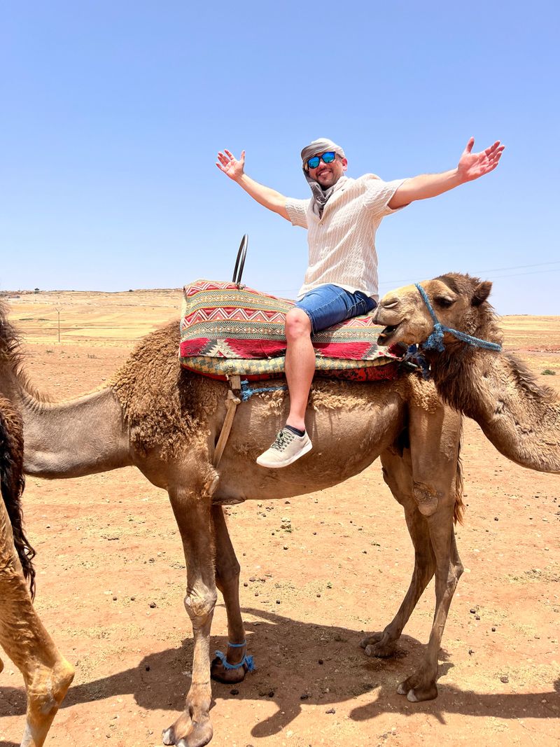 Mid adult latin tourist male wearing a turban enjoying a camel ride in the Moroccan Sahara desert with arms outstretched, celebrating freedom and adventure
