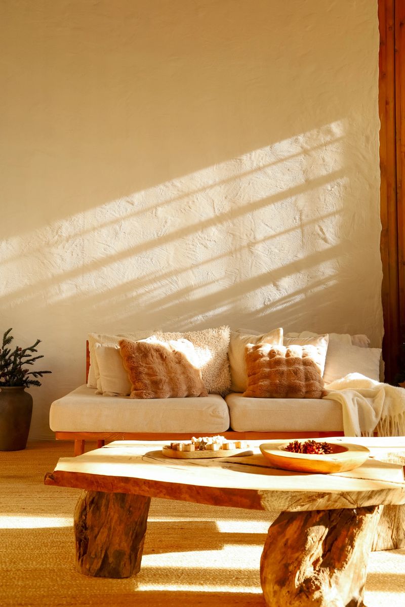 A cozy living room features a large white couch decorated with soft pillows. A wooden coffee table holds snacks while natural light streams through the window creating a welcoming atmosphere.