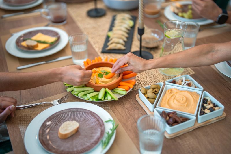 Two hands, one reaching for hummus, are seen at a dinner party table featuring fresh vegetables, dips, and snacks. The setting is warm and inviting, with a variety of dishes spread across the wooden table.