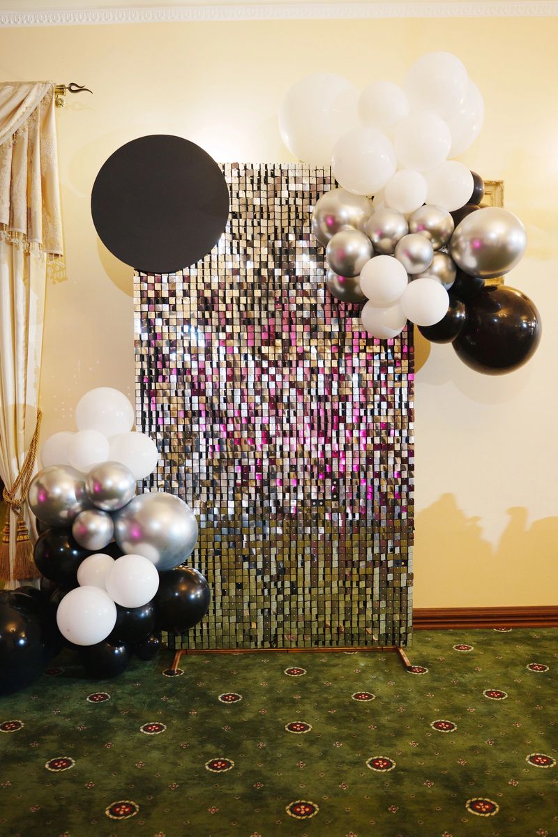 A vibrant balloon display with black, white, and silver elements is arranged beside a shimmering backdrop in a decorated hall for a festive event.