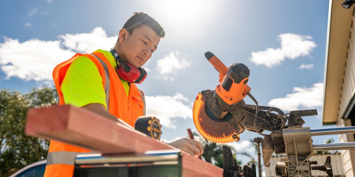 Worker in safety gear marking wood near a power saw.