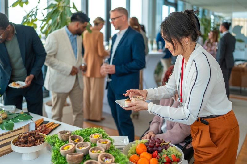 A group of adults are seen selecting food at a business meeting. The setting is a modern conference room with a buffet table featuring a variety of dishes. Attendees are engaged and networking.