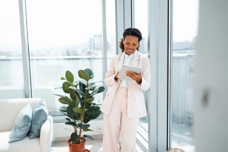 Stylish professional woman standing by a window in a modern office, engaging with a tablet device. The setting features natural light, stylish decor, and an atmosphere of productivity and professionalism.