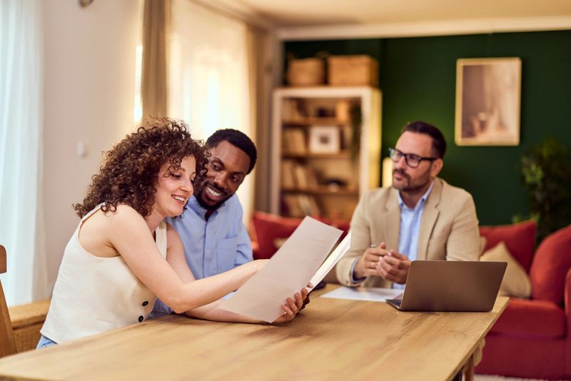 A young couple chats with an insurance or real estate agent over documents in a warm, homey office.