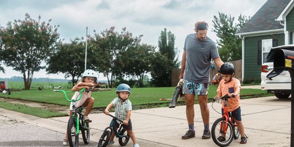 A man and three children with bikes on a suburban street, all wearing helmets.