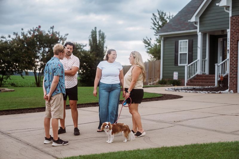 A small group of adults chats warmly in a suburban driveway with a dog at their feet, enjoying a relaxed moment together.