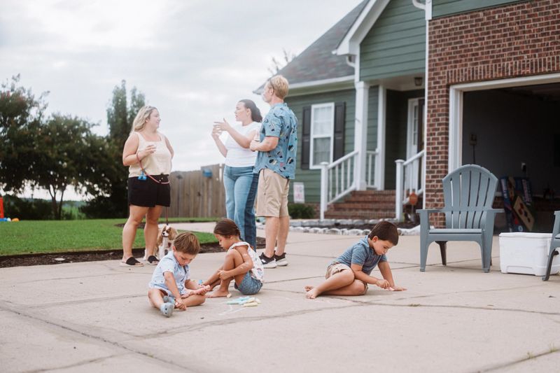 Kids sit on the driveway making chalk drawings while nearby adults talk casually during a relaxed neighborhood gathering.