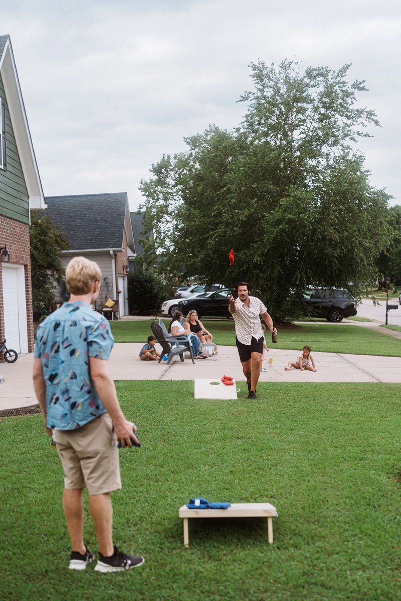Two men play a friendly game of cornhole in the front yard of a suburban home while a mix of kids and adults visit and relax in the nearby driveway.