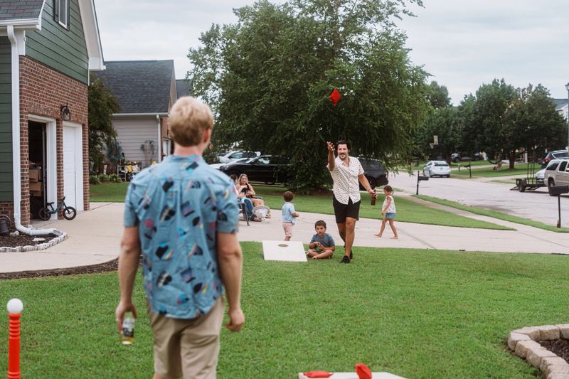 Two men play a friendly game of cornhole in the front yard of a suburban home while a mix of kids and adults visit and relax in the nearby driveway.