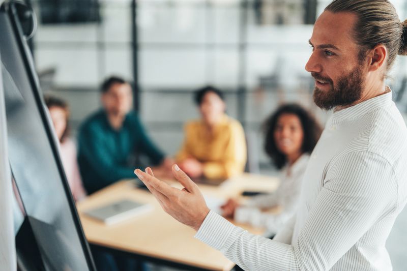 Businessman standing in front of flipchart and showing ideas to coworkers, teamwork on business strategy. Smiling male mentor or coach make flipchart presentation for diverse employees, excited presenter present project on whiteboard, coworkers at casual briefing. Young businessman presenting business strategy on white board to colleague in the office