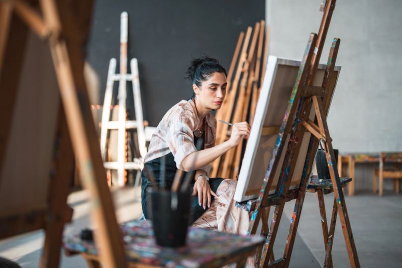 A young Middle Eastern female artist works intently on a canvas in a brightly lit artistic studio, surrounded by wooden easels and art supplies.
