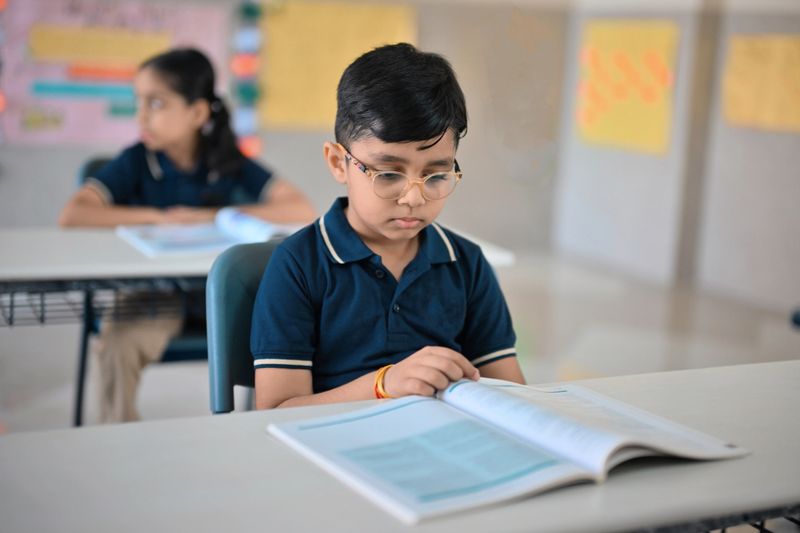 A young boy attentively reads a textbook while seated at his desk in a brightly lit primary school classroom. The student is dressed in uniform, appears focused and engaged with his learning material.