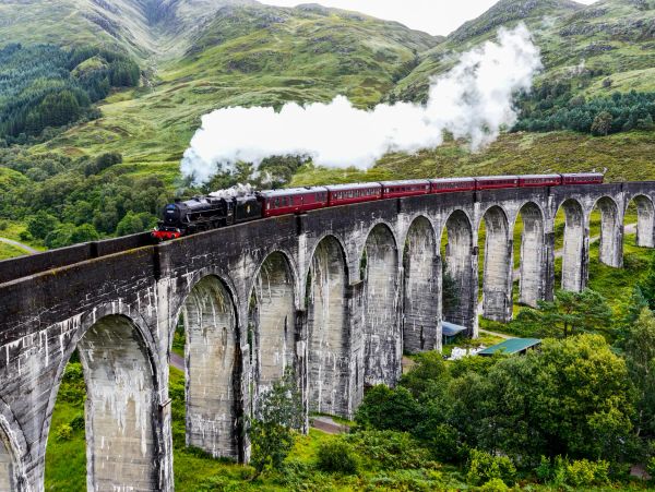 Steam train crossing a stone viaduct in a green mountainous area.