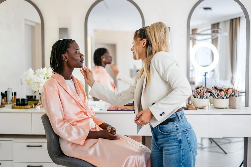 Young black woman getting her makeup done by a female beautician at the beauty salon. Female beautician erasing mistakes on her client's lips with a q-tip. Close-up.