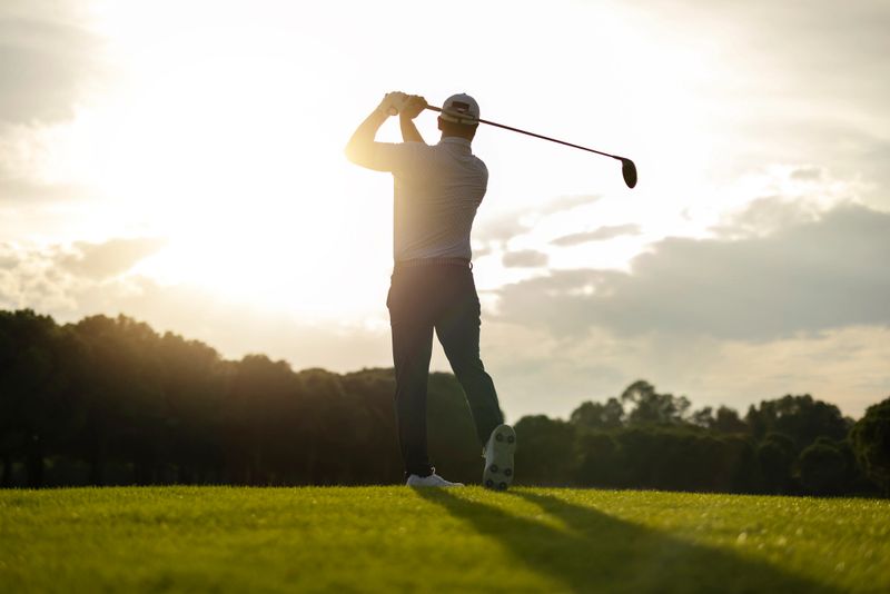 Silhouette of a golfer preparing to swing at sunrise on a bright fairway. Warm backlight and a calm outdoor atmosphere make the image suitable for sports, golf lifestyle, motivation, and leisure themes.