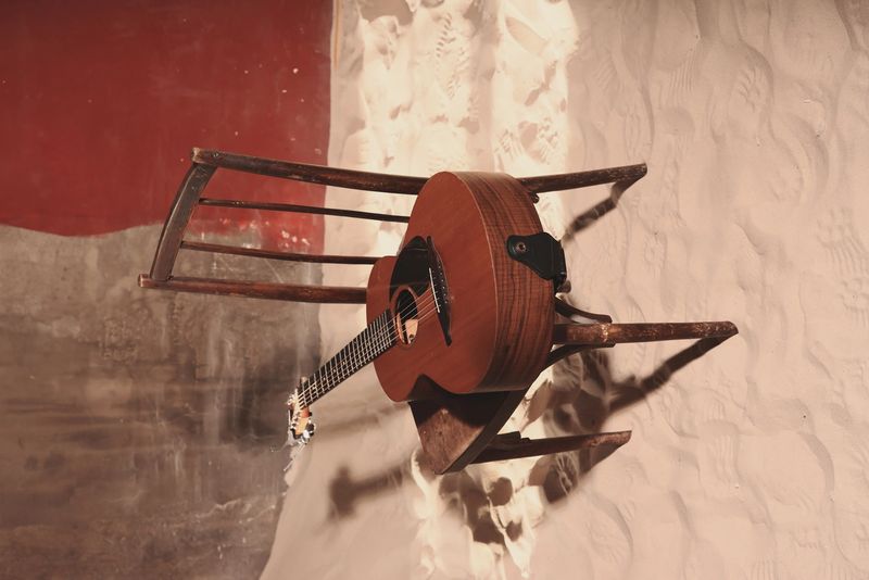 acoustic guitar on wooden chair in textured room, warm sunlight and sharp shadow across frets, rustic walls and minimal setup
