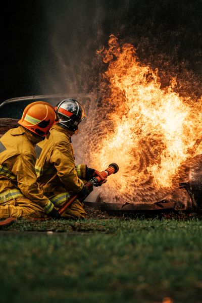 Two firefighters in yellow gear extinguishing a large fire with a hose.