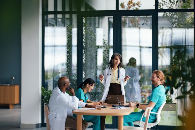 A diverse medical team gathers around a wooden table in a bright, modern hospital or clinic space. The standing doctor explains points while teammates listen, with laptops and tablets open nearby.