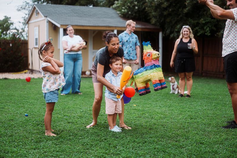 A Eurasian woman helps her young son grip a plastic bat as he prepares to take his turn at hitting a colorful piñata at his big sister's birthday party.