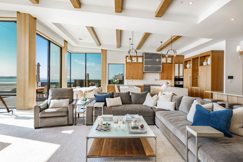 Image shows the interior of a luxurious mountain modern living room with large floor to ceiling windows with a view of the ocean over an expansive patio with a swimming pool and a hot tub. It is a beautiful blue sky sunny summer day. The room has a contemporary design with a stone fireplace wood cabinetry and panoramic views. A large grey sectional sofa modern light fixtures and a telescope are visible in the space.