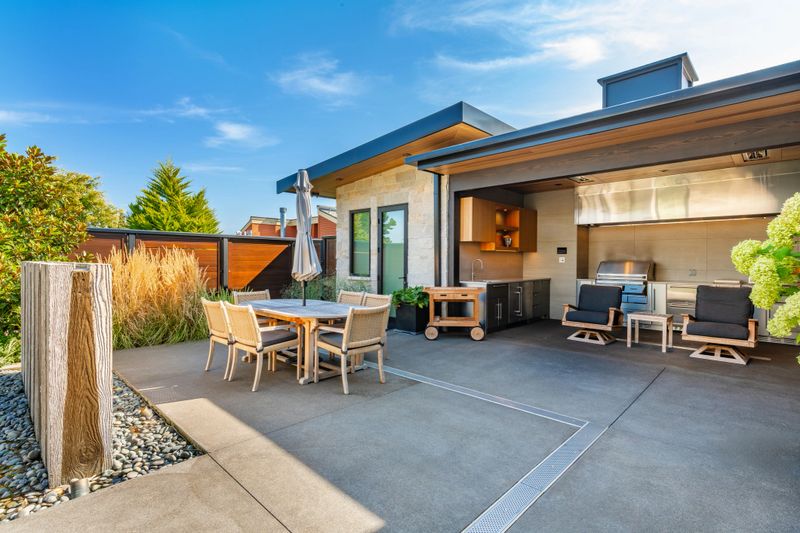 Exterior photograph of a mountain modern home residence a bright blue sky summer day and the house has an expansive ocean front deck with a swimming pool and a hot tub the tide is low and there are coastal views we also see an enclosed outdoor living area with stainless steel cabinets cooking facilities dining area wooden furniture the front facade of the home shows a flat roof and two large garage doors with glass windows the home has stone and wood cladding waterfront deck leads to modern living room through floor to ceiling glass doors