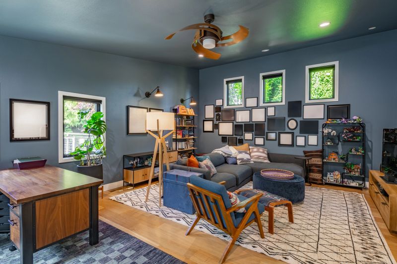 This is a large modern home interior with a distinct bar area and adjacent living room characterized by warm wood finishes and a contemporary design. Striking wooden paneling and shelving in the wet bar area custom cabinetry shows open shelving wine cubbies drawers and lower cabinets. Living room interior shows blue accent walls a grey sectional sofa an armchair and patterned rug creating a cozy and inviting atmosphere. Rooms show a fireplace and a loft space. The space has a bohemian aspect and lots of decor and accents that show a family lives here. Natural setting