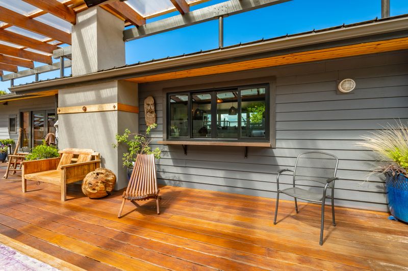 Image displays a large modern outdoor living space, specifically a covered patio and deck area. The space features extensive wooden decking and a slatted wooden roof with translucent panels for light and to shield from rain. A full outdoor kitchen is visible, including a built in pizza oven, stainless steel grill and a smoker. There are distinct dining and lounging areas with a large wooden dining table a long bench and comfortable outdoor seating. A concrete retaining wall forms the back of the patio, and string lights are installed across the ceiling for ambiance. There are several potted plants, ferns and flowers. The photograph was taking on a bright sunny summer day with blue sky and filtered sunlight.